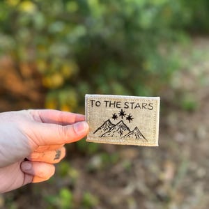May include: A beige fabric patch with the text "TO THE STARS" and a black embroidered design of mountains and stars.
