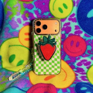 May include: A clear phone case with a green and white checkered pattern and a red strawberry pop-socket. The phone case has an orange back and a clear strap with beads. The background is a colorful fleece fabric with smiley faces.