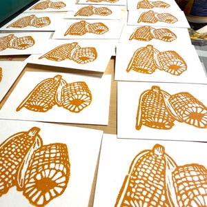 May include: A collection of handmade art prints featuring a detailed illustration of two corn cobs. The prints are in a warm orange hue, creating a rustic and inviting aesthetic. The prints are arranged on a light-colored surface.