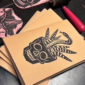 May include: A stack of brown paper notebooks with black ink illustrations of a skull with a snake and plants growing out of the top. The notebooks are stacked on a black surface.