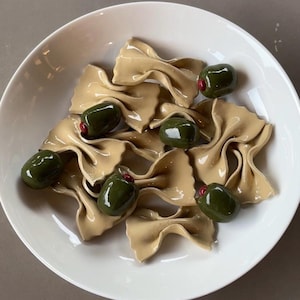 May include: A white ceramic bowl filled with tan-colored farfalle pasta and green olives. The olives have a red center. The pasta is arranged in a decorative pattern. The bowl is on a neutral surface.