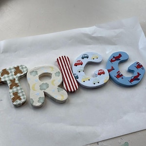 May include: Wooden letters spelling out the name "TRACE". The letters are painted with various patterns, including teddy bears, clouds, cars, airplanes, and stripes. The letters are in shades of blue, red, white, and beige.
