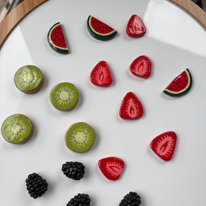 May include: A collection of colorful fruit-shaped magnets, including watermelon slices, kiwi rounds, strawberry halves, and blackberries. The magnets are arranged on a white surface, with a wooden frame in the background. The magnets are likely for a refrigerator or a magnetic board.