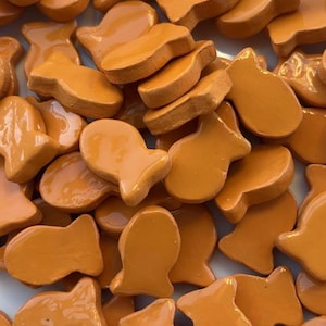 May include: A close-up of a pile of orange, fish-shaped biscuits. The biscuits have a glossy finish and are scattered across the frame. The biscuits are a uniform orange colour.