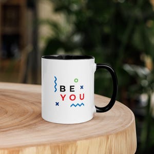 May include: White ceramic mug with a black handle and interior. The mug features the words "BE YOU" in black, red, and blue, with colorful geometric shapes. The mug is sitting on a wooden surface.