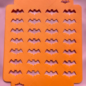 May include: Orange rectangular Halloween-themed craft stencil with bat cutouts. The stencil has a textured surface and a series of bat shapes cut out in rows and columns. The stencil is designed for crafting and creating Halloween decorations.