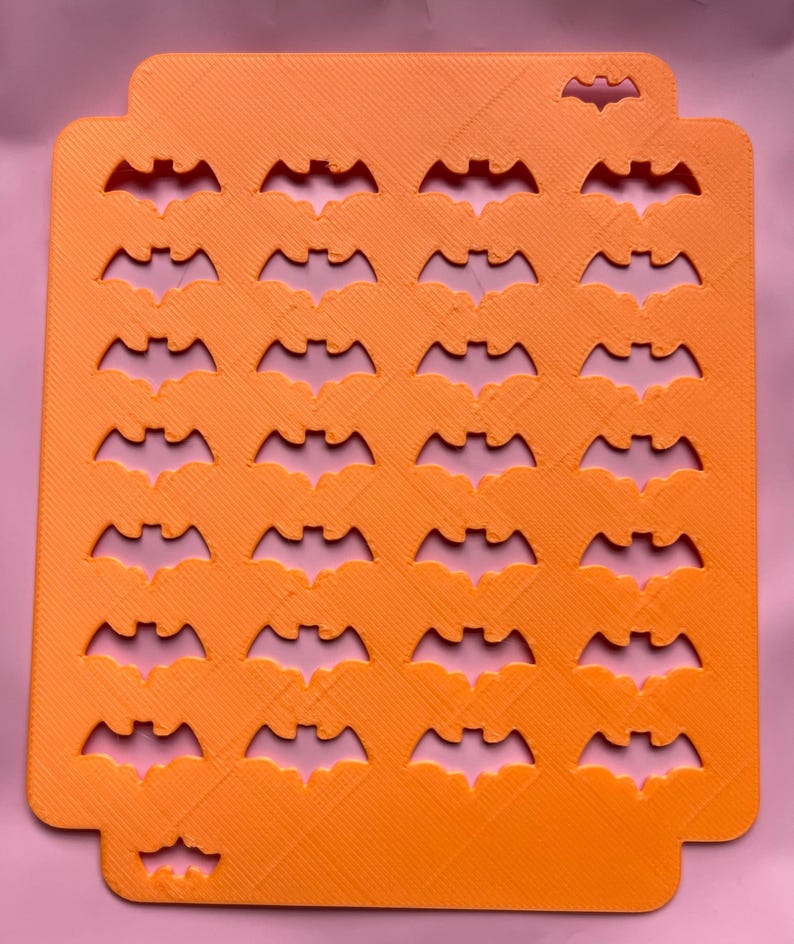May include: Orange rectangular Halloween-themed craft stencil with bat cutouts. The stencil has a textured surface and a series of bat shapes cut out in rows and columns. The stencil is designed for crafting and creating Halloween decorations.