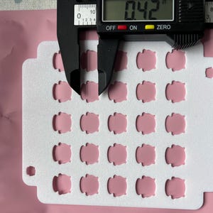 May include: A close-up shot shows a white card with 25 small, pink, rounded-square shapes. A digital caliper measures the thickness at 0.42 inches. The background is a pink, sealed plastic bag.