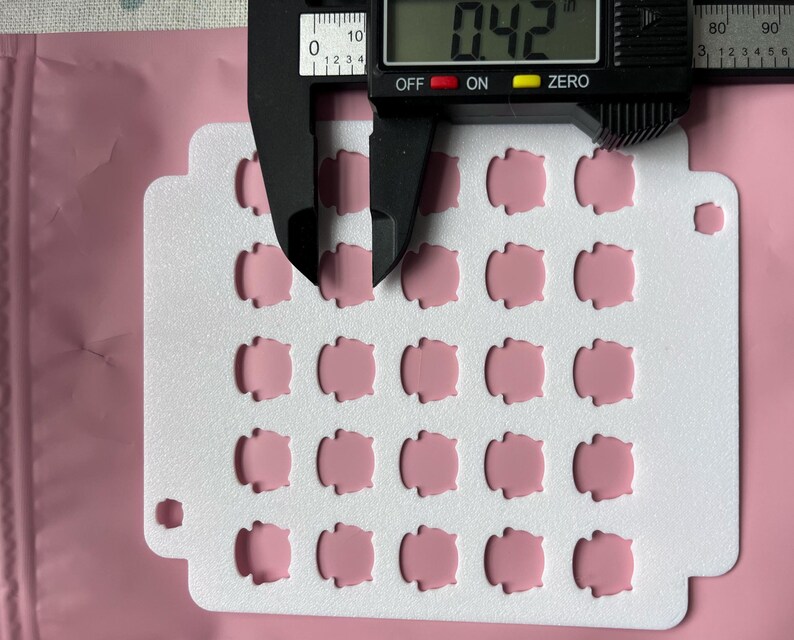 May include: A close-up shot shows a white card with 25 small, pink, rounded-square shapes. A digital caliper measures the thickness at 0.42 inches. The background is a pink, sealed plastic bag.