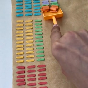 May include: Rows of colorful, small, rectangular objects in blue, yellow, green, and red are arranged on a brown surface. An orange scoop holds more of the objects. A hand is holding the scoop.