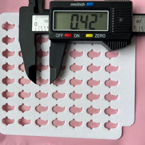 May include: A sheet of pink, fish-shaped embellishments, each measuring 0.42 inches. A digital caliper displays the measurement. The embellishments are arranged on a white, die-cut card, set against a pink background.