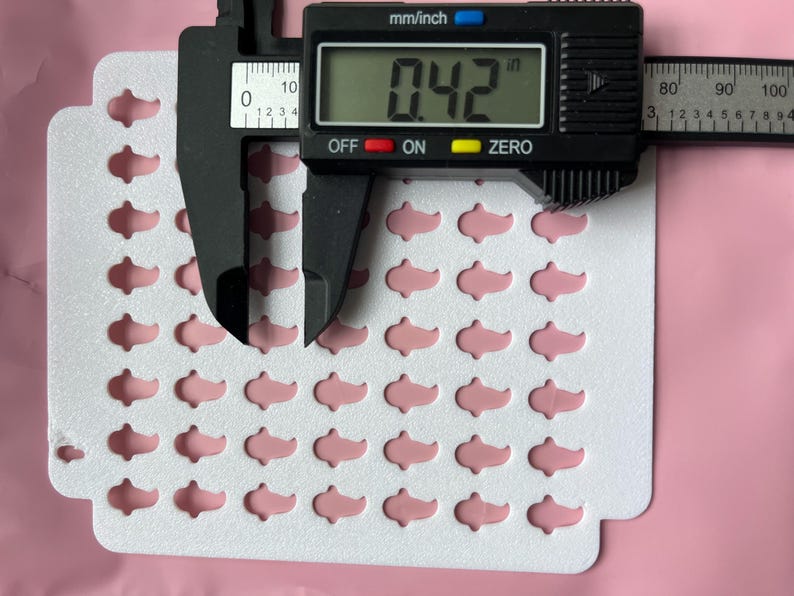 May include: A sheet of pink, fish-shaped embellishments, each measuring 0.42 inches. A digital caliper displays the measurement. The embellishments are arranged on a white, die-cut card, set against a pink background.