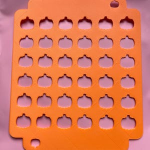 May include: Orange plastic pumpkin-shaped cookie cutter with 30 cutouts. The cutter has a rectangular shape with rounded corners and a small hole at the top for hanging. The pumpkin shapes are evenly spaced throughout the cutter.