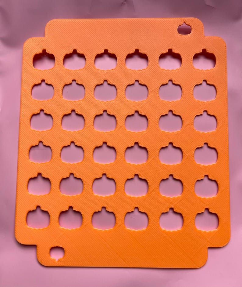 May include: Orange plastic pumpkin-shaped cookie cutter with 30 cutouts. The cutter has a rectangular shape with rounded corners and a small hole at the top for hanging. The pumpkin shapes are evenly spaced throughout the cutter.