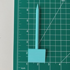 May include: A light blue 3D-printed stylus, approximately 6 inches long, with a rectangular base. The stylus is positioned on a green cutting mat with inch measurements. The mat has a grid pattern and angle markings.