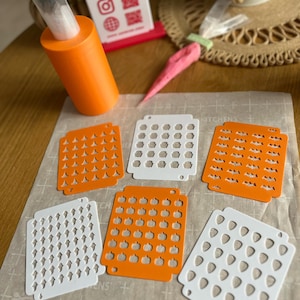 May include: An orange icing bag holder with a clear plastic bag inside, and several white and orange stencils with pumpkin and bat cutouts. A pink and green icing carrot is also visible.