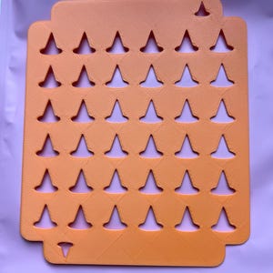 May include: An orange, rectangular organizer with a series of inverted triangle cutouts. The organizer has a textured surface and a small cutout at the top and bottom. The cutouts are evenly spaced and create a grid-like pattern.