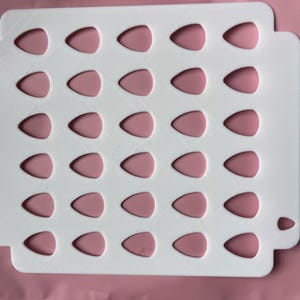 May include: A white, rectangular tray with a series of teardrop-shaped cutouts, each filled with a pink, teardrop-shaped insert. The tray has rounded corners and a textured surface. The background is a solid pink color.