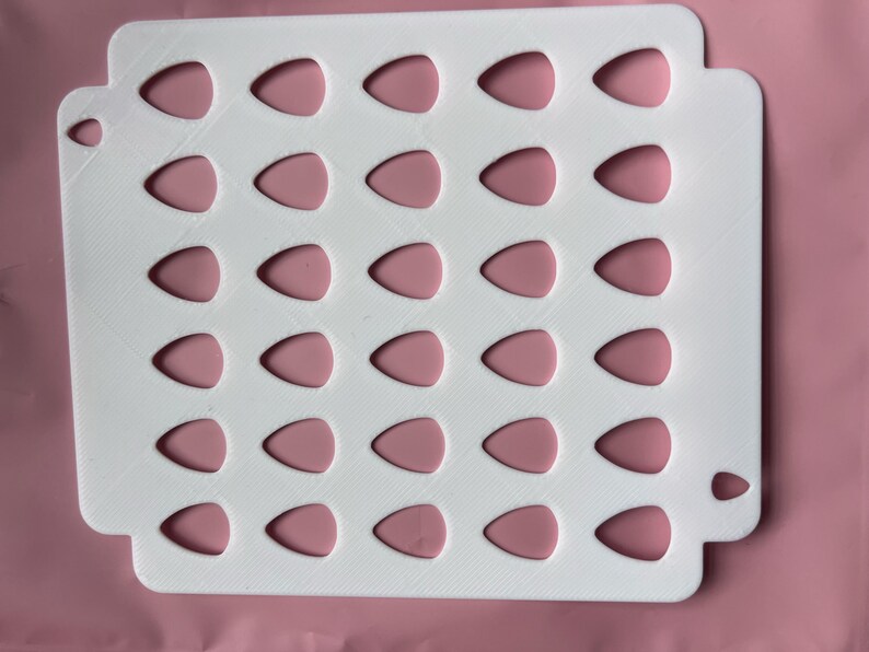 May include: A white, rectangular tray with a series of teardrop-shaped cutouts, each filled with a pink, teardrop-shaped insert. The tray has rounded corners and a textured surface. The background is a solid pink color.