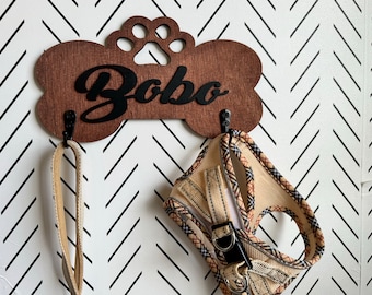 May include: A wooden dog bone-shaped hook with the name "Bobo" engraved on it. The hook has two metal hooks for hanging leashes or collars. A tan and black patterned dog harness is hanging from one of the hooks.