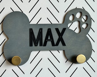 May include: Gray wooden dog bone shaped sign with the name "MAX" in black lettering and a paw print cutout. The sign has two gold hooks on the sides.