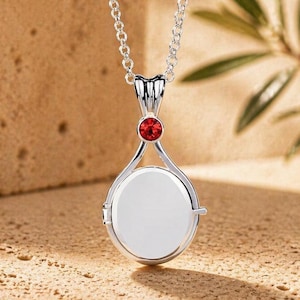 May include: A silver pendant necklace featuring a circular, white centre. A small, red gemstone sits above the pendant. The necklace is suspended from a fine chain. The background is a neutral tan colour.
