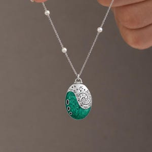 Peut inclure: Collier en argent avec un pendentif ovale vert orné d'un motif en spirale blanc et de trois petits points noirs. La chaîne comporte de petites perles blanches. Le collier est tenu par une main.