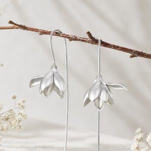 Puede incluir: Un par de pendientes colgantes plateados. Cada pendiente presenta un diseño en forma de flor suspendido de un alambre largo y delgado. Se exhiben sobre una superficie de piedra, con una ramita y pequeñas flores blancas.