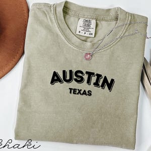 May include: Khaki-coloured t-shirt with the word "AUSTIN" in bold black letters above "TEXAS". The shirt has a Comfort Colors tag and a silver necklace with a pendant. A brown hat is partially visible in the upper left corner.