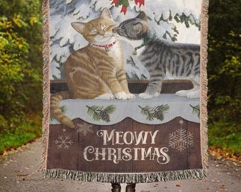 May include: A woven throw blanket with a winter scene featuring two cats kissing under a snowy evergreen tree. The text "MEOWY CHRISTMAS" is printed on a wooden sign below the cats.