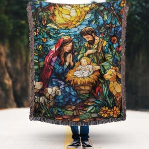 Könnte beinhalten: Eine gewebte Decke mit einer Darstellung der Geburt Christi im Stil eines Glasfensters. Das Kunstwerk zeigt Maria, Josef und das Jesuskind in warmen Farben wie Blau, Rot und Gelb. Die Decke hat einen Fransenrand.