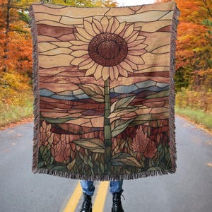 May include: A woven tapestry throw blanket featuring a stained glass style sunflower with a brown, yellow, and green color scheme. The sunflower is set against a background of a landscape with brown, green, and blue hues.