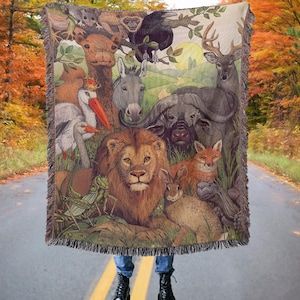 May include: Woven throw blanket featuring a detailed illustration of various animals, including a lion, deer, and fox. The artwork showcases a vibrant color palette with a natural theme. The blanket has a fringed edge and is displayed against a road backdrop.