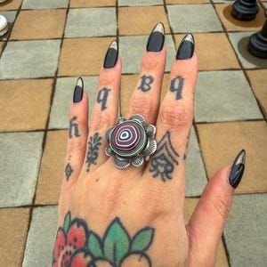 May include: A silver ring with a flower design and a central stone with red, purple, and white stripes. The hand has black nail polish with gold tips and tattoos. Chess pieces are in the background.