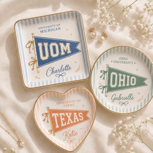 May include: Three decorative trinket dishes in square, round, and heart shapes. Each dish features a pennant design with a university name and a name. The square dish has a blue pennant, the round dish has a green pennant, and the heart dish has an orange pennant.
