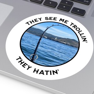 May include: A white circle sticker with black text that reads "They See Me Trollin' They Hatin'". The sticker features a photo of a fishing rod with a line in the water. The background is a blue sky and water with a coastline in the distance.