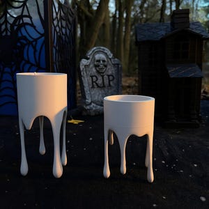 Puede incluir: Dos candelabros blancos con un diseño de cera goteando. Los soportes son cilíndricos con tres gotas de cera que forman las patas. Una lápida con "RIP" y una pequeña casa negra están en el fondo, sugiriendo un tema de Halloween.