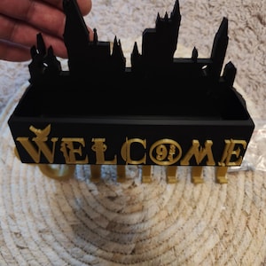 Könnte beinhalten: Schwarzer Schreibtisch-Organizer mit einer Silhouette einer Schloss-Skyline. Das Wort "WELCOME" ist in goldfarbenen Buchstaben auf der Vorderseite zu sehen, mit der Nummer "9 3/4" in der Mitte. Der Organizer ist aus dunklem Material.