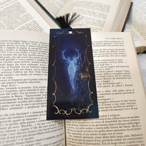 Può includere: Un segnalibro nero e oro con un'illustrazione blu e bianca di un cervo in piedi di fronte a un edificio. Il segnalibro ha il testo "A Darker Than Black Mystery" in basso.