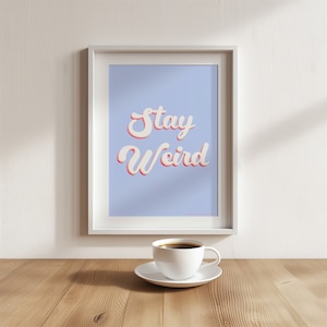Può includere: Una stampa incorniciata bianca con uno sfondo blu chiaro e il testo "Stay Weird" in un font retrò bianco con un contorno rosa. Una tazza da caffè bianca e un piattino sono in primo piano.