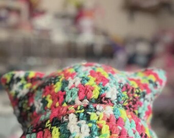 May include: A colorful, handmade crochet hat with cat ears. The hat features a vibrant mix of pink, green, yellow, and white yarn. The hat is worn low, partially obscuring the eyes and black-framed glasses.