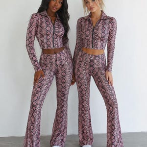 Op de afbeelding: Een tweedelige set bestaande uit een crop top met lange mouwen, kraag en ritssluiting aan de voorzijde, en een bijpassende high-waisted broek met uitlopende pijpen. Beide stukken hebben een roze en zwarte slangenprint. Gecombineerd met witte sneakers.
