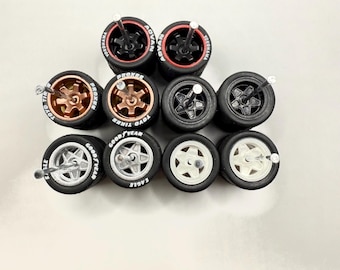 May include: A collection of miniature model car wheels. The wheels feature various designs, including black, bronze, and silver rims with black tires. Some tires have white lettering with brand names like "ADVAN" and "GOOD YEAR".