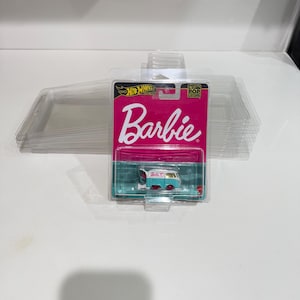 May include: A Hot Wheels Barbie Pop Culture die-cast van in a clear plastic package. The van is light blue and gold with red wheels. The packaging has a pink background with the Barbie logo.