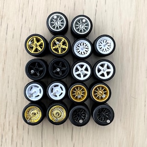 May include: A collection of 18 miniature car wheels in various styles. The wheels are black, white, and gold with different spoke designs. Some wheels have a chrome finish.