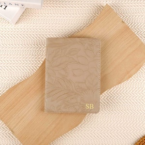 May include: A beige passport holder with an embossed floral design and gold initials "SB". The cover is made of a smooth material and is set on a wooden board and a woven surface.