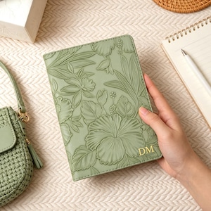 May include: A sage green journal with embossed floral designs and gold initials. The journal is held by a person's hand. A matching green handbag, a notepad with a pen, and a woven coaster are also in the image.