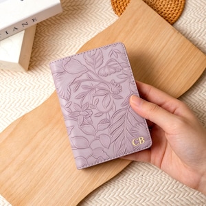 May include: A lavender-colored passport holder with an embossed floral design. The cover features a detailed pattern of flowers and leaves. Gold initials "CB" are visible on the lower right corner. The holder is held on a wooden cutting board.