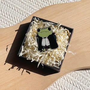 Könnte beinhalten: Ein grüner Frosch-Emaille-Pin mit schwarzem Umhang und weißem Hemd, präsentiert in einer schwarzen Geschenkbox mit geschreddertem Papier. Der Frosch hat große Augen und einen kleinen Mund. Der Pin liegt auf einer Holzoberfläche.