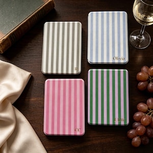 May include: Four rectangular metal tins featuring vertical stripes in gray, light blue, pink, and green. Each tin is personalized with gold lettering: R, Olivia, CC, and LEW. The tins are arranged on a dark surface alongside a book, wine glass, and grapes.